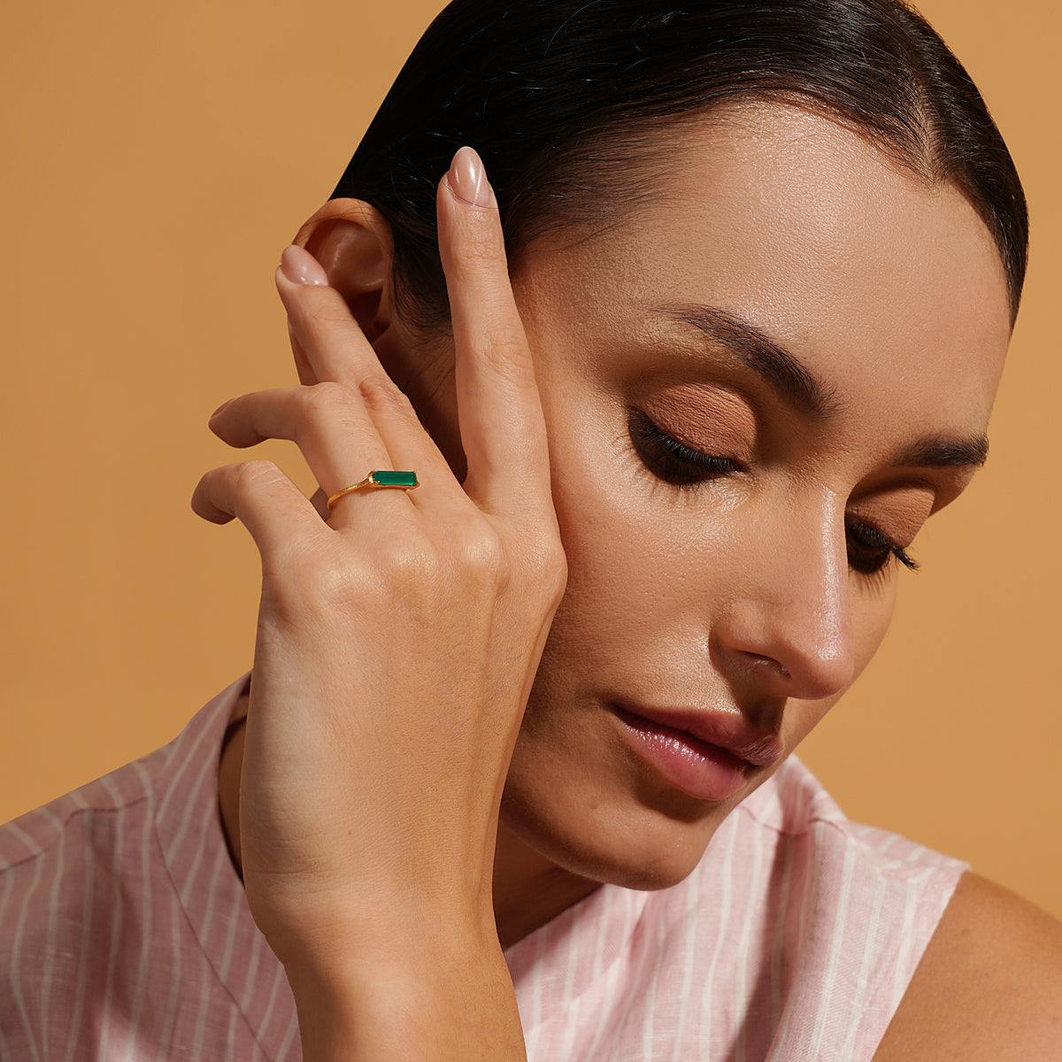 Close-up of a hand wearing a delicate gold ring set with a slim emerald-green baguette stone. Warm natural light highlights the stone's rich color and the ring's minimalist design against a softly blurred background.
